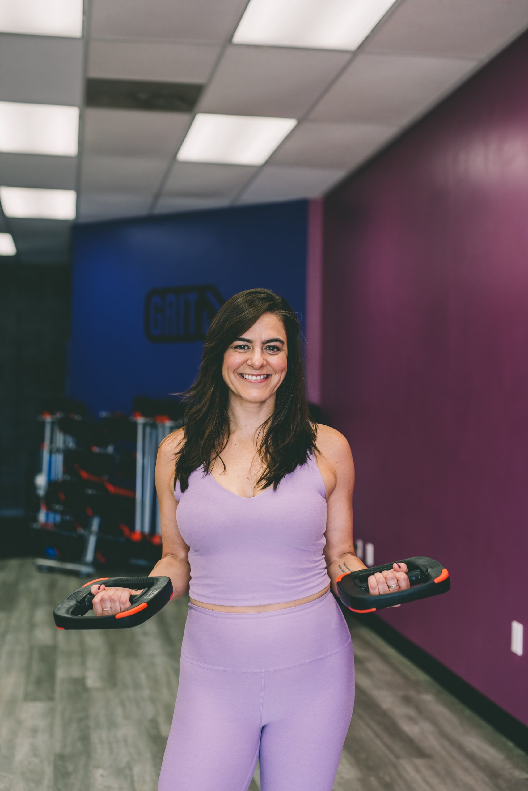 Melissa Banford está de pie con un atuendo de entrenamiento de lavanda en el gimnasio, sosteniendo pesas con ambas manos y sonriendo a la cámara.