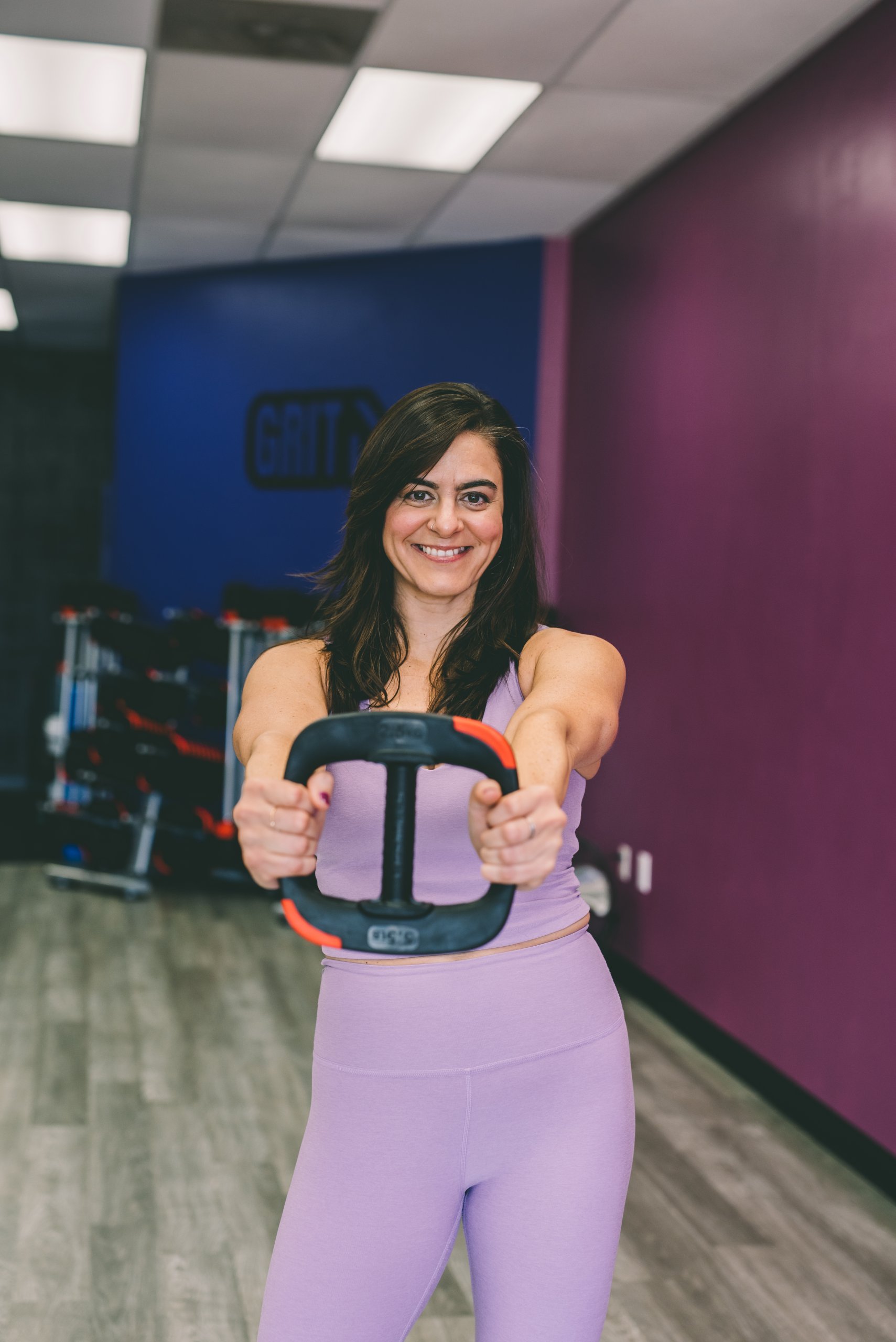 Vestida con un atuendo de entrenamiento de lavanda, Melissa Banford sostiene una retaguardia con las manos en un gimnasio dibujando una pared morada.