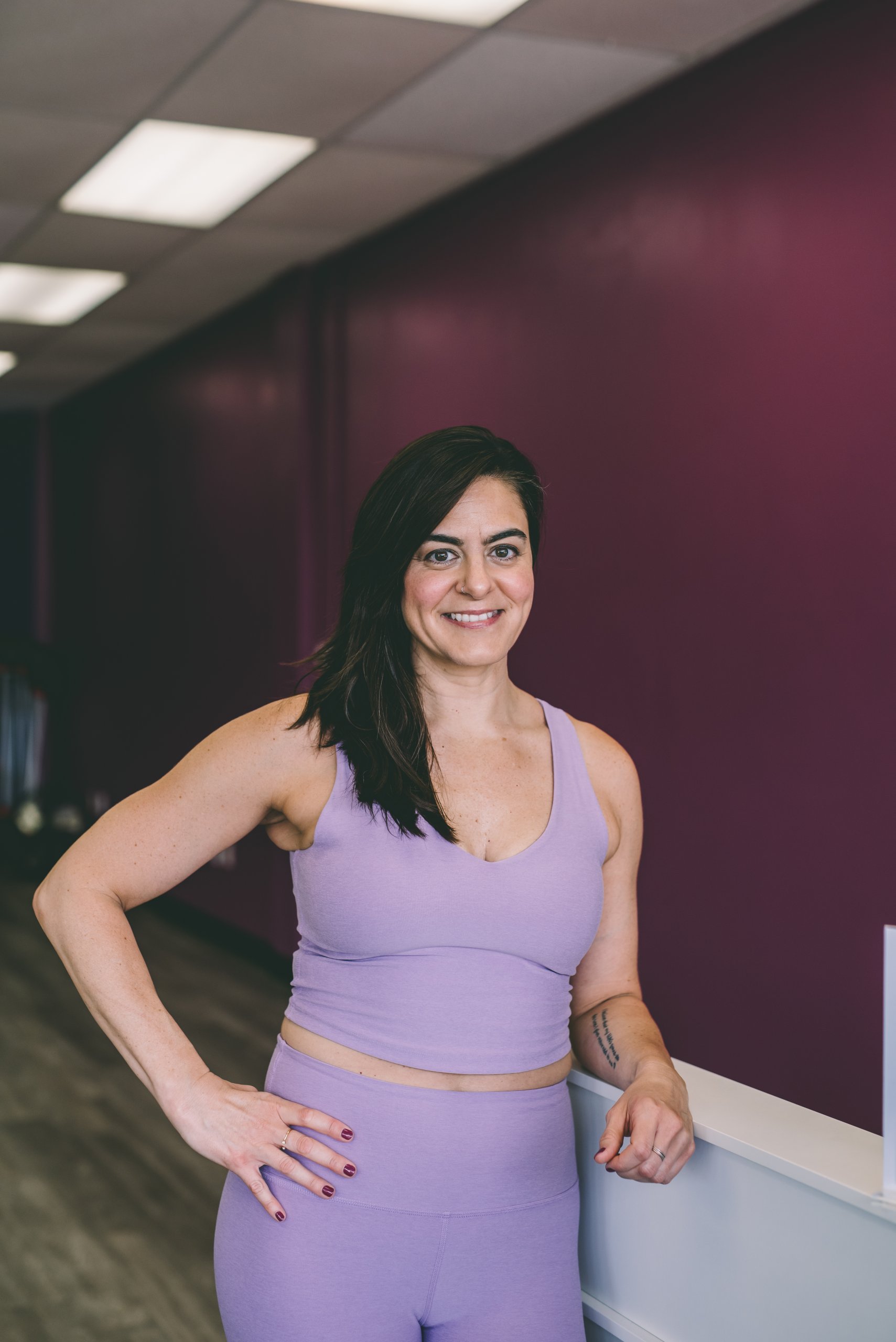 Melissa Banford se encuentra en el interior contra una pared púrpura, sonriendo con su sujetador deportivo de lavanda y leggings con una mano.