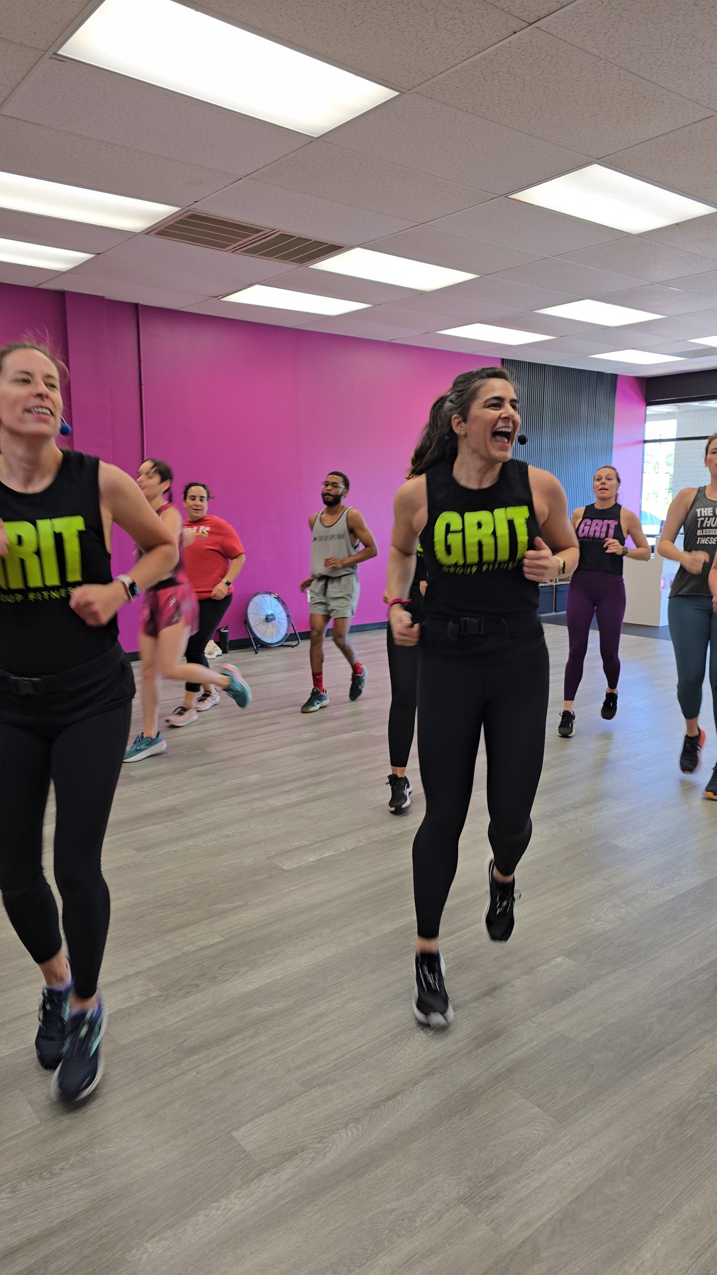 Un grupo de personas que asisten a la clase de fitness de la cara, dirigida por dos mujeres negras, es "Arena" Camisa: una de ellas es Melissa Bamford. La habitación tiene paredes rosadas y blancas y grandes ventanas.