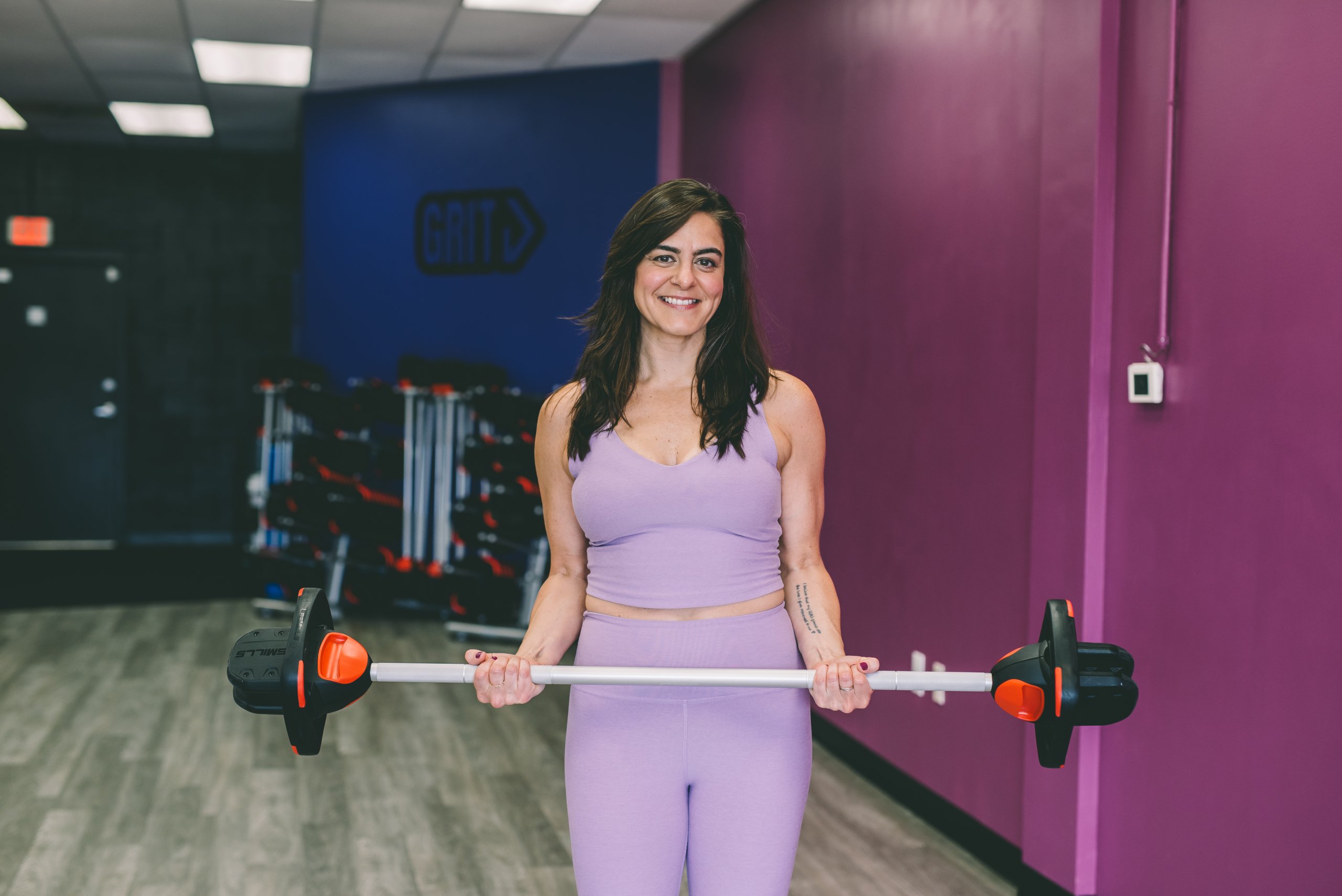 Melissa Bamford levanta la barra y sonríe con el atuendo de entrenamiento de lavanda de su fe, un gimnasio brillantemente iluminado con paredes moradas.