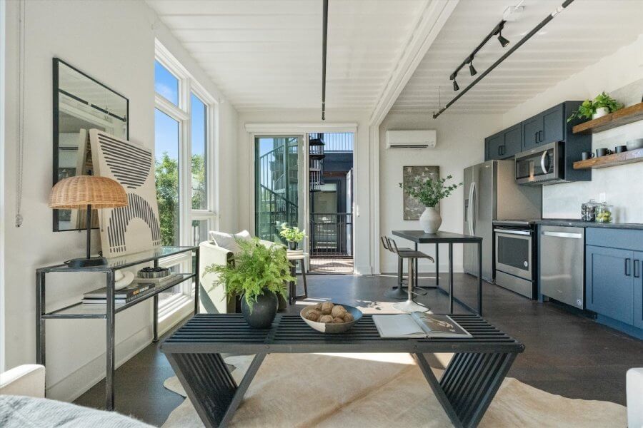 Modern open-concept living and kitchen area at 83 Freight in Wedgewood-Houston, Nashville, with blue cabinets, large windows, a black coffee table, green plants, minimalist decor, and abundant natural light.