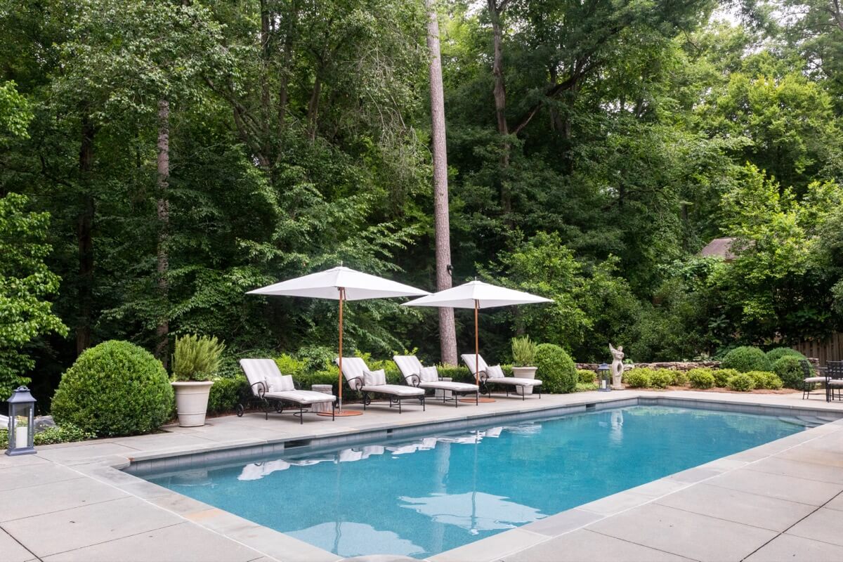 A rectangular outdoor swimming pool with lounge chairs and white umbrellas, surrounded by lush greenery and trees, offers a serene retreat in the heart of Buckhead, Atlanta—perfect for relaxing at home.