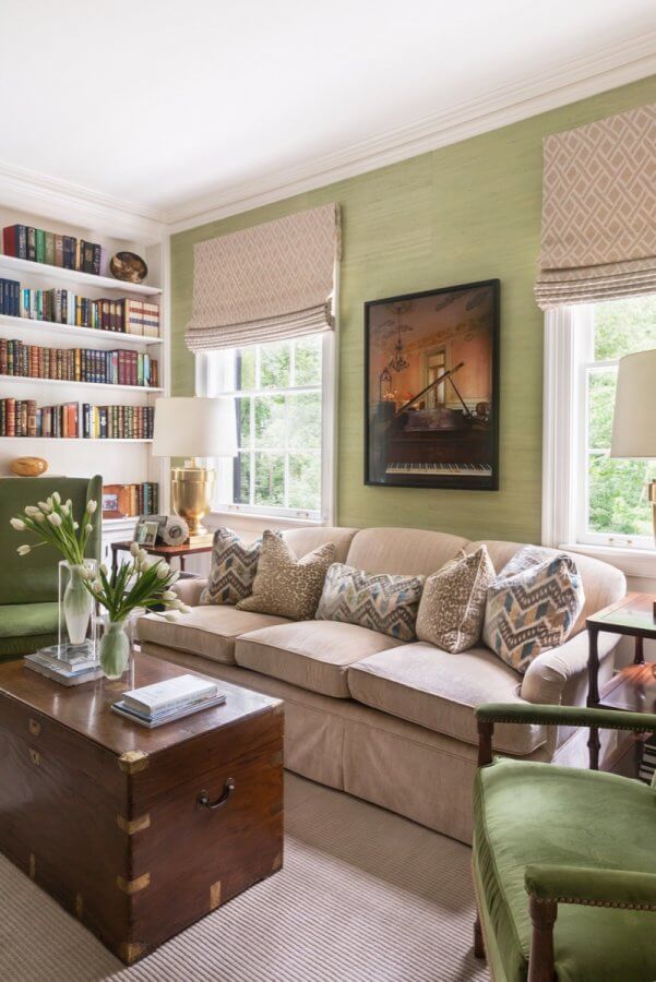 A cozy Atlanta home living room with a beige sofa, patterned pillows, green walls, bookshelves, large windows with Roman shades, a wooden chest as a coffee table, and green accent chairs.