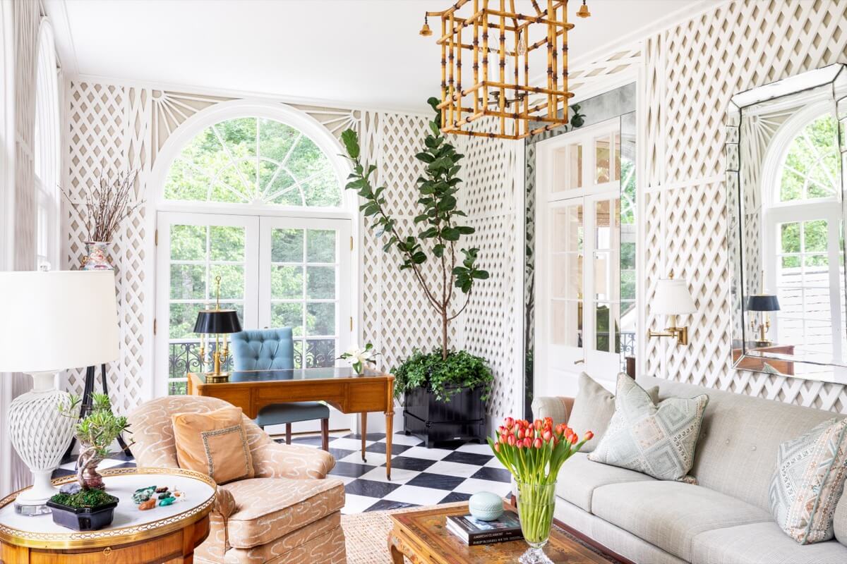Bright sunroom in a Buckhead home with large arched windows, patterned walls, checkered floor, desk, sofa, potted plant, and gold chandelier; tulips on the wooden coffee table add charm to this inviting Atlanta space.
