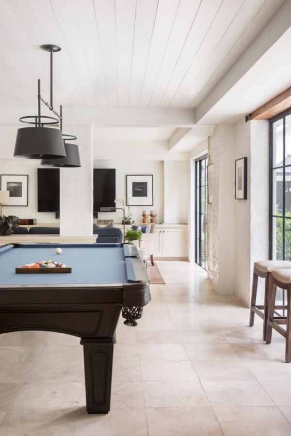 A modern Buckhead game room with a black pool table, bar stools by large windows, light tile flooring, and minimalist wall deco.