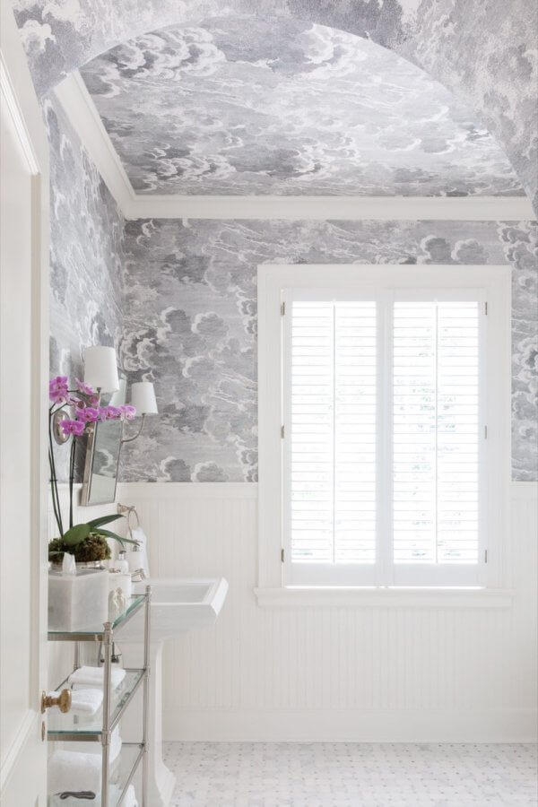 Bright Buckhead bathroom with patterned wallpaper on walls and ceiling, white window shutters, pedestal sink, glass shelf, and a purple orchid in a pot add charm to this inviting Atlanta home.
