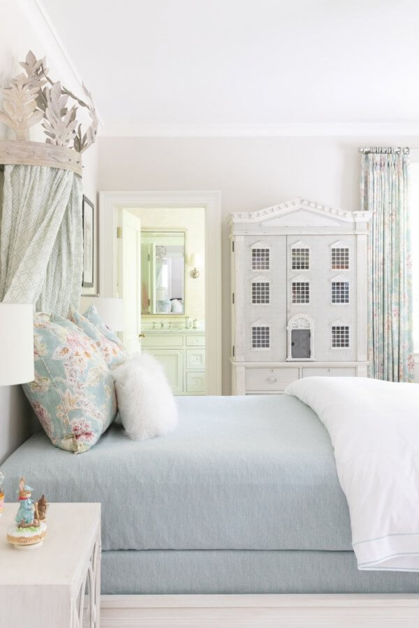 A pastel-colored bedroom in Buckhead, Atlanta, features a light blue bed, floral pillows, a white nightstand with figurines, light curtains, and a large dollhouse-shaped cabinet.