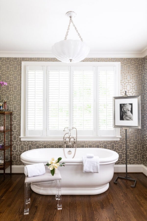 A white freestanding bathtub sits in front of large shuttered windows in this Buckhead home, with a towel and flower on a clear bench, and a framed photo on a stand beside patterned wallpaper.