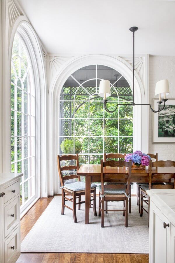 A bright Buckhead dining area with large arched windows, a wooden table and chairs, a chandelier, and a vase of purple and blue flowers on the table creates an inviting Atlanta home ambiance.