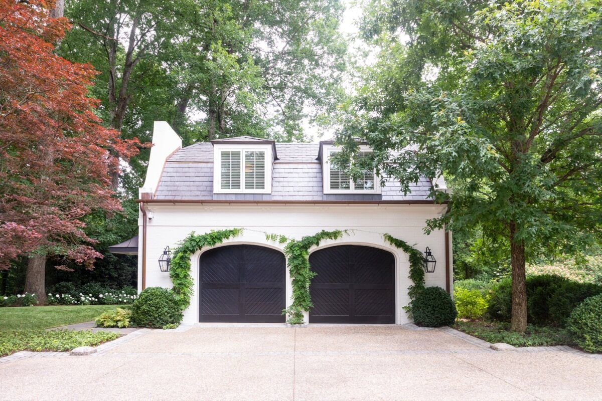A detached two-car garage with dark wooden doors, white exterior walls, green ivy, and dormer windows is surrounded by trees and landscaped greenery—an inviting home feature in Atlanta’s Buckhead neighborhood.