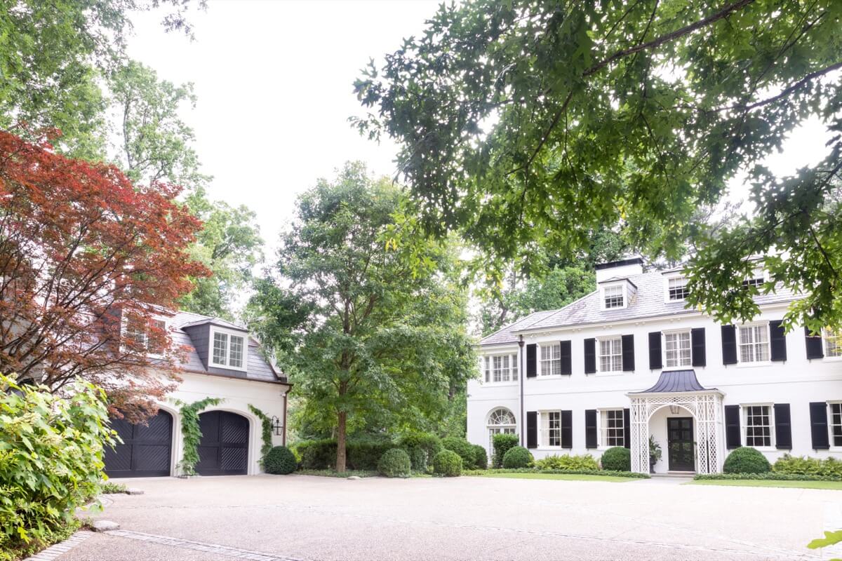 Large white colonial-style Atlanta Buckhead home with black shutters, columns, and a prominent front door; a detached garage and trees are visible in the foreground.