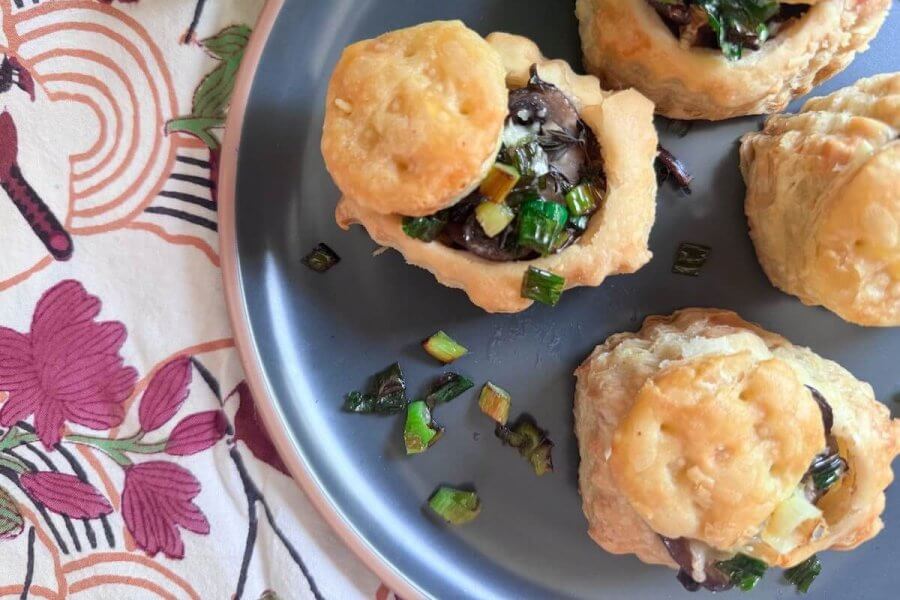 Three puff pastry bites filled with a mushroom and herb mixture are arranged on a blue-gray plate, with chopped green onions scattered nearby—a delicious addition to your collection of savory puff pastry recipes.