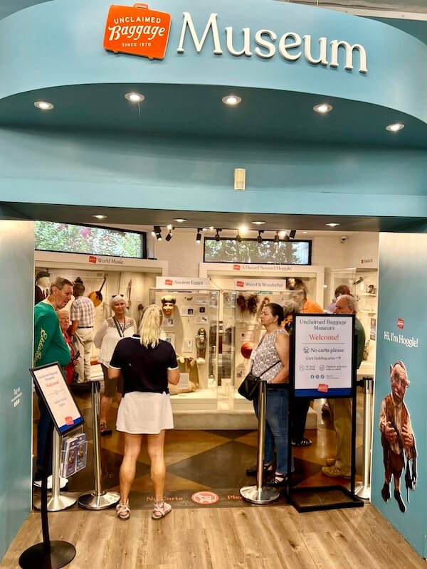 A group of people stand at the entrance of the Unclaimed Baggage Museum, known for its connection to discount stores, looking at exhibits and signs under a blue archway.