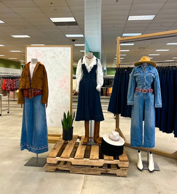 Three mannequins display Western-inspired outfits with denim pieces inside a retail clothing store, reminiscent of styles often found in discount stores; shelves of jeans and a whiteboard are visible in the background.