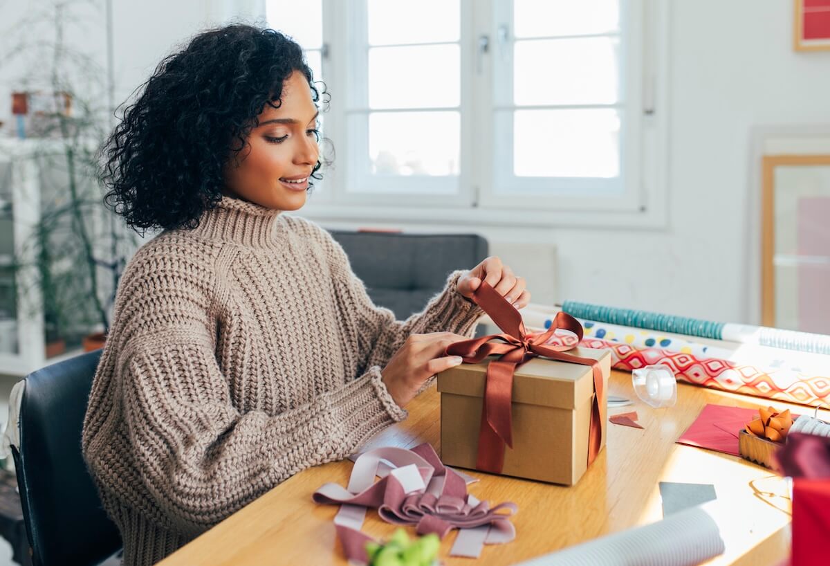 The New Rules of Financial Etiquette: Splitting the Bill, Venmo Requests & More - 3 A woman sits at a table tying a red ribbon on a wrapped gift box, surrounded by wrapping paper—her thoughtful touch reflecting modern money manners and the art of financial etiquette in today's gifting traditions.