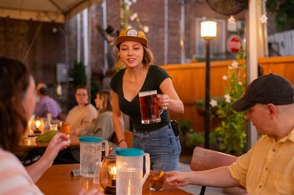 A server hands a glass of beer to a seated customer at an outdoor Nashville restaurant in the evening, where other patrons enjoy food in the background.