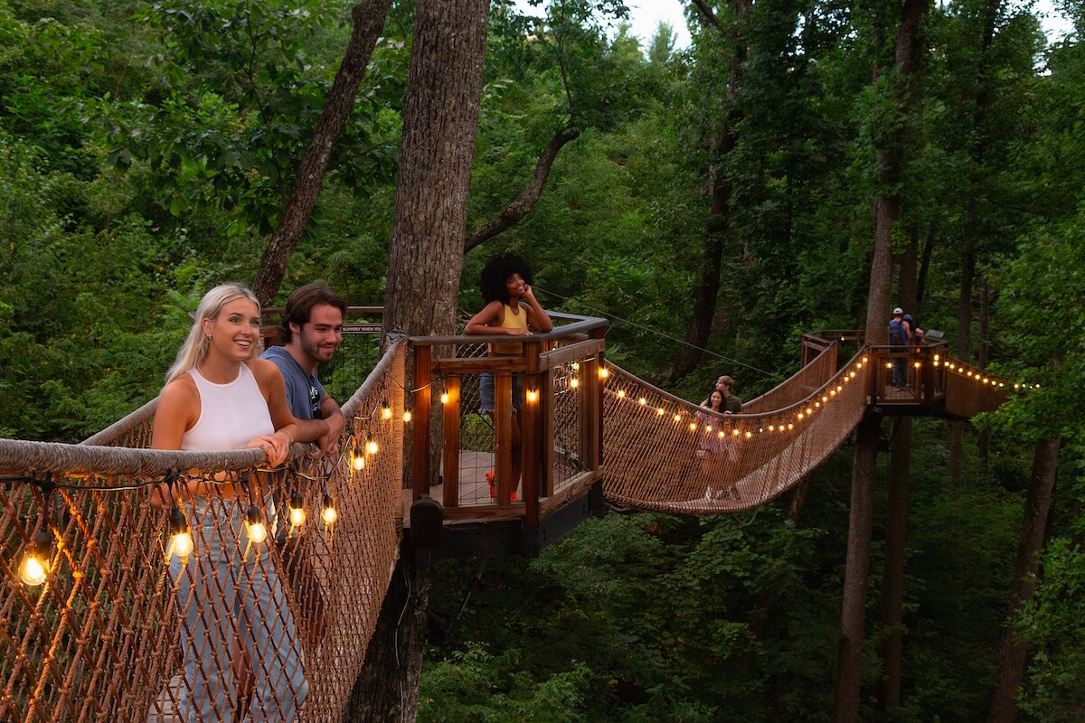 5 Beloved Tennessee Destinations for Adventure Lovers - 3 People stand on a wooden suspension bridge strung with lights, surrounded by dense green forest. Some lean on the railing, while others walk along the bridge, enjoying one of Tennessee outdoor escapes and true adventure towns.