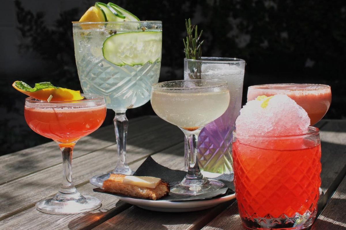 Six colorful cocktails in various glassware on a wooden table, garnished with fruit, herbs, and a pastry—perfect for enjoying Birmingham happy hours as sunlight casts playful shadows.