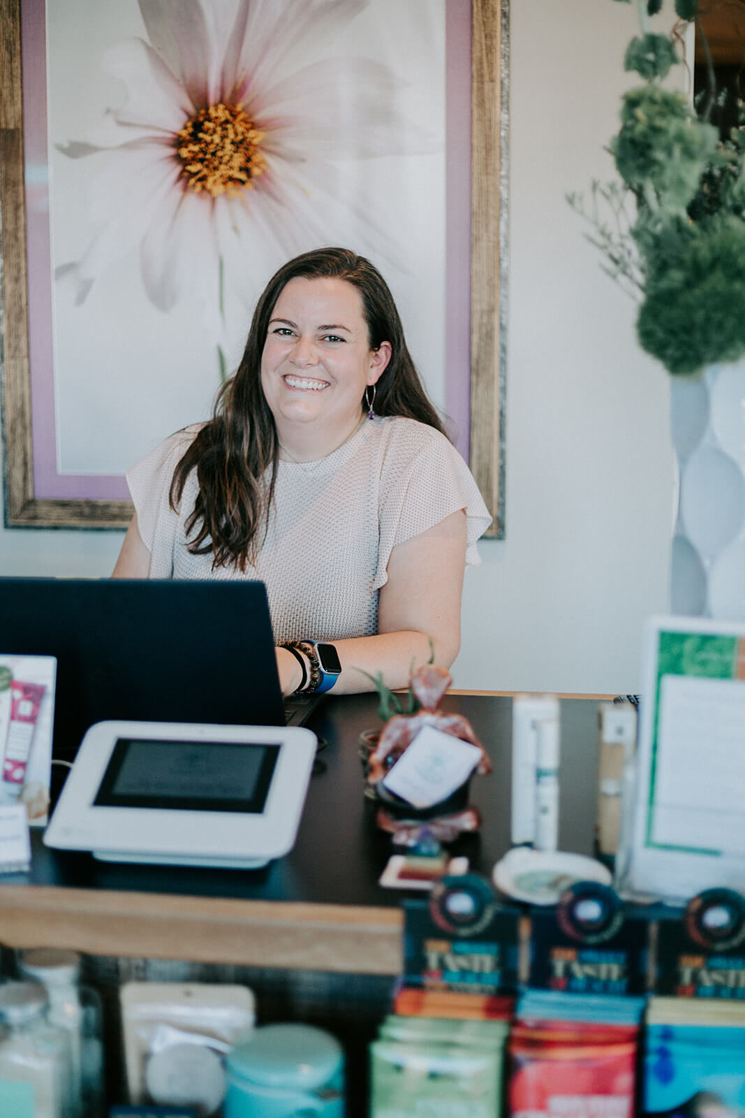 She’s Behind Two of Birmingham’s Most Beloved Day Spas - 2 Jill Copeland sits behind a reception desk at one of Birmingham’s top day spas, smiling by her computer, with various products displayed in the foreground and a large flower photo on the wall behind her.