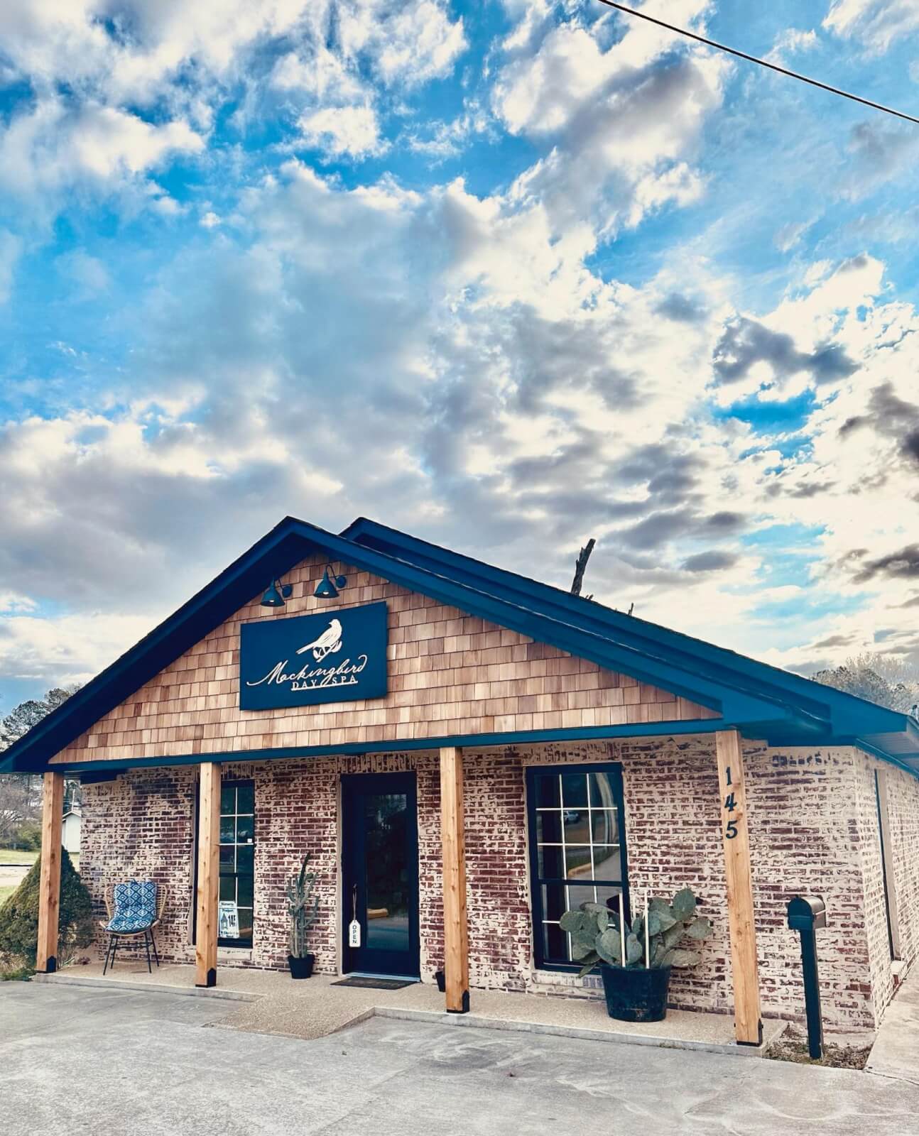 She’s Behind Two of Birmingham’s Most Beloved Day Spas - 4 A small brick building with wood columns and a sign above the entrance that reads "Mockingbird Day Spa" under a partly cloudy sky.