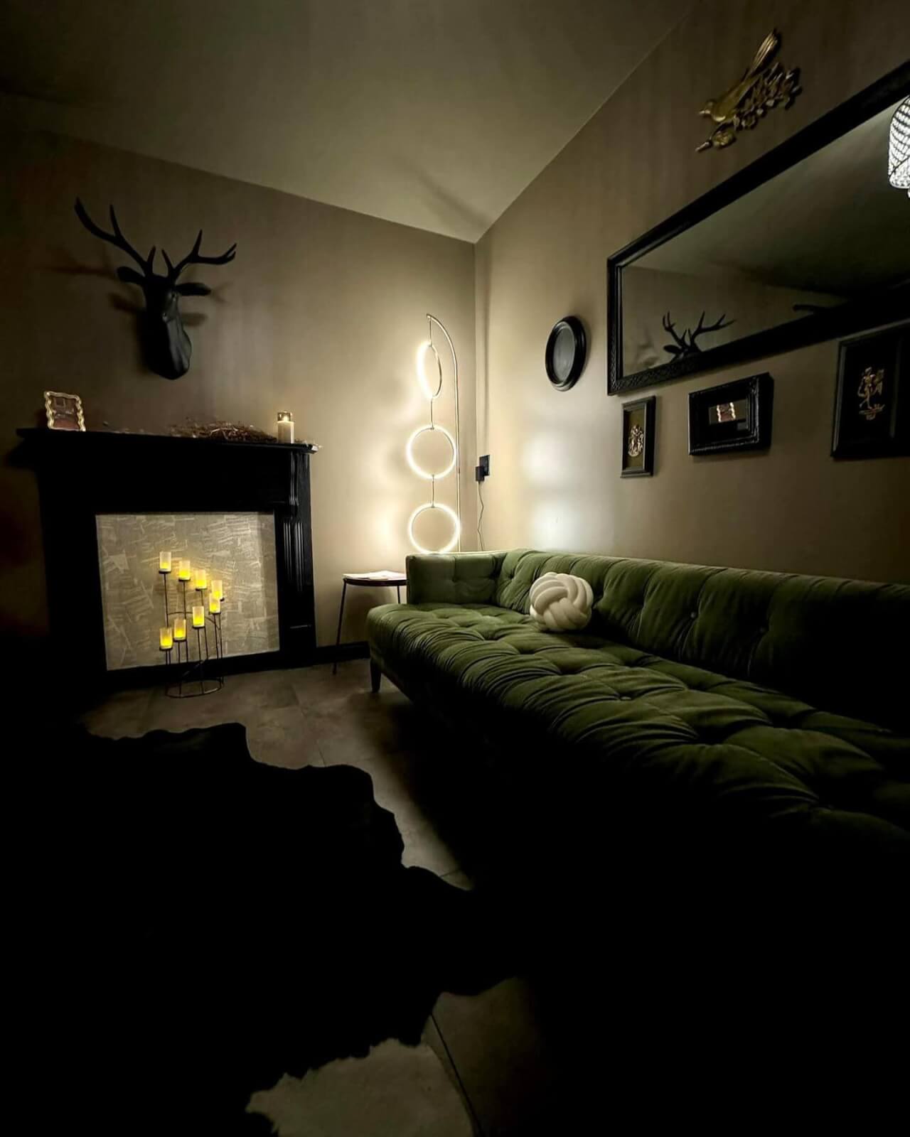 She’s Behind Two of Birmingham’s Most Beloved Day Spas - 6 The dimly lit spa room at Mockingbird Day Spa features a plush green sofa, wall art, a faux deer head, a mirror, a modern floor lamp, and LED candles in a fireplace.