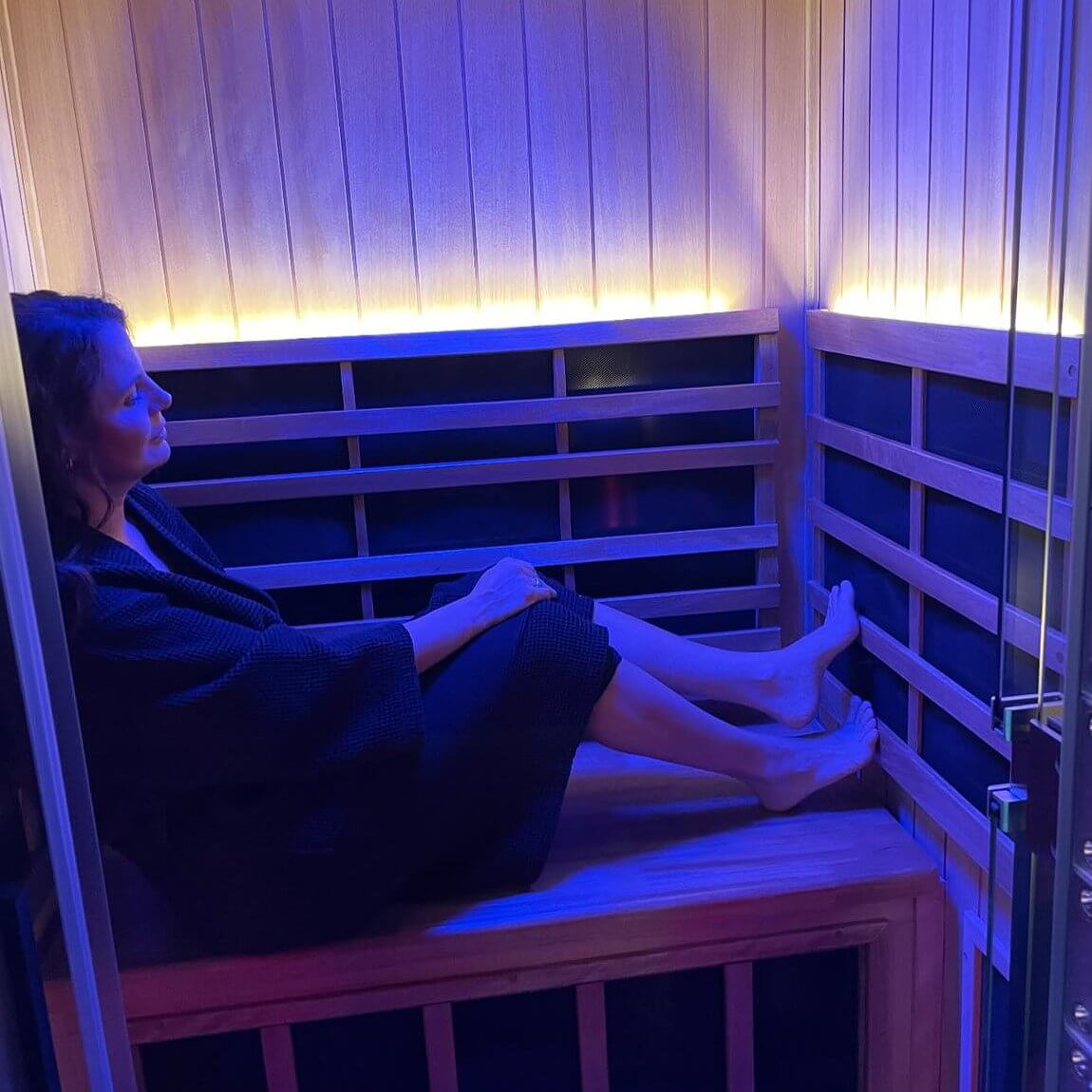 She’s Behind Two of Birmingham’s Most Beloved Day Spas - 8 A person leaning back and relaxing in a dark robe sits inside a small, wooden infrared sauna with blue and yellow lighting at Mockingbird Day Spa.