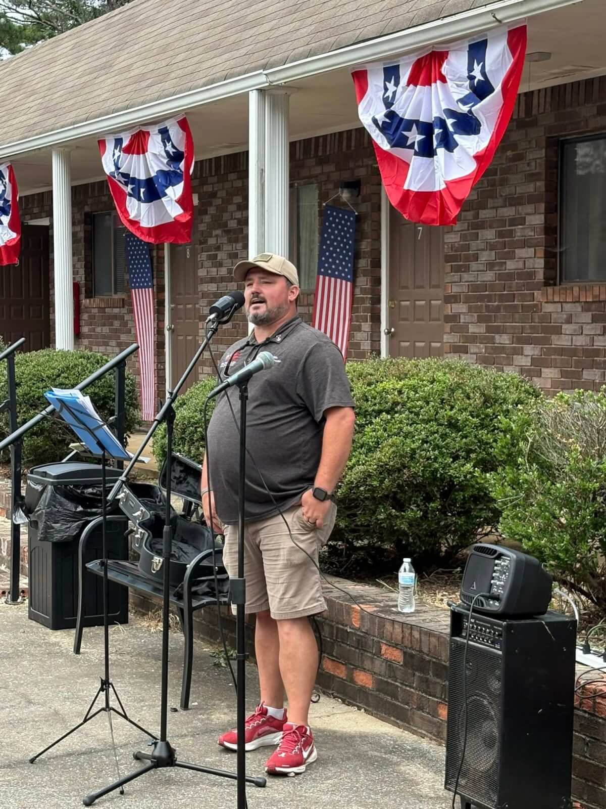 How 'Three Hots and a Cot' is Supporting Birmingham Veterans - 3 Executive Director of Three Hots and a Cot Jay Gibbs stands and sings into a microphone outdoors, with patriotic banners and American flags decorating the new Oneonta Veteran's Housing Center behind him.