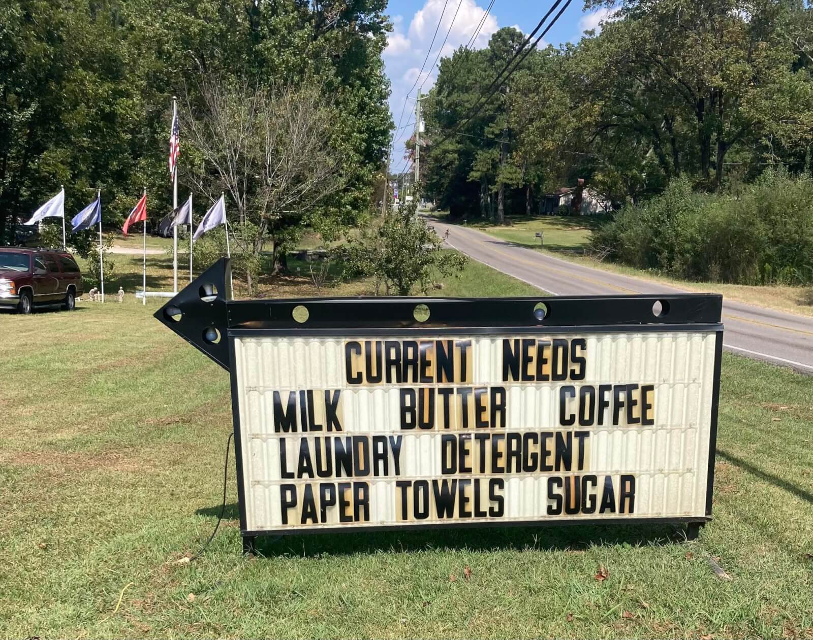 How 'Three Hots and a Cot' is Supporting Birmingham Veterans - 6 An outdoor sign for Birmingham nonprofit Three Hots and a Cot lists current needs: milk, butter, coffee, laundry detergent, paper towels, and sugar. Flags and trees stand in the background beside a road.