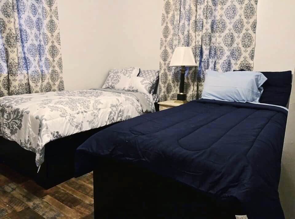 How 'Three Hots and a Cot' is Supporting Birmingham Veterans - 2 Two twin beds, one in blue and the other in a gray and white pattern, sit side by side with a nightstand and lamp between them—creating a welcoming space featured in our Nonprofit Spotlight on Three Hots and a Cot, a Birmingham nonprofit.