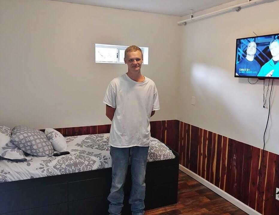 How 'Three Hots and a Cot' is Supporting Birmingham Veterans - 4 A person stands next to a bed in a small room with wood paneling, a window, and a wall-mounted TV at the Birmingham Nonprofit Three Hots and a Cot.
