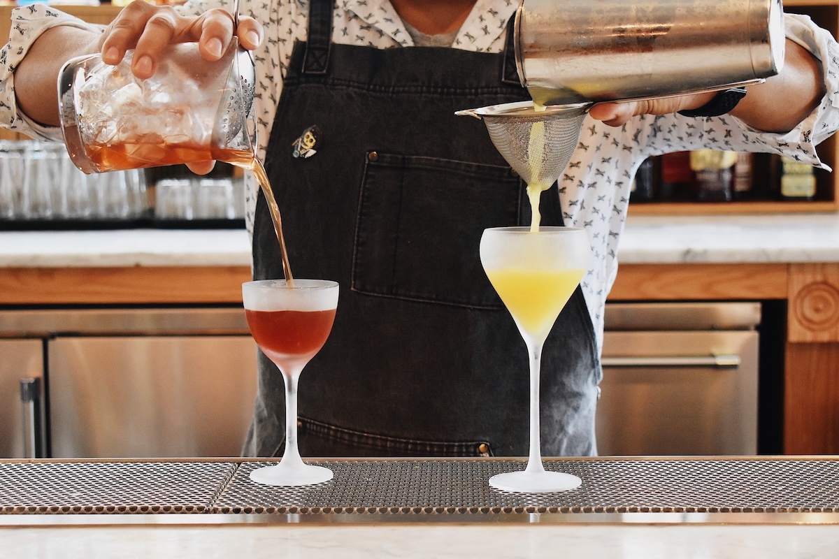 A bartender pours two different cocktails into chilled glasses, using a strainer for the yellow drink—setting the perfect scene for a Memphis happy hour.