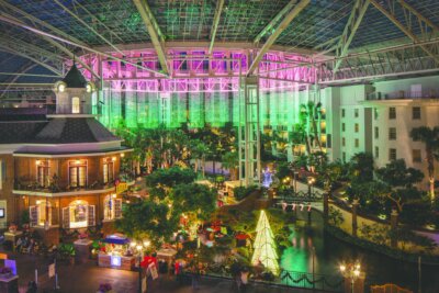 An indoor hotel atrium with a glass ceiling, glowing pink and green lights, a canal, and a courtyard featuring a lit Christmas tree, a popular destination for Nashville November events.