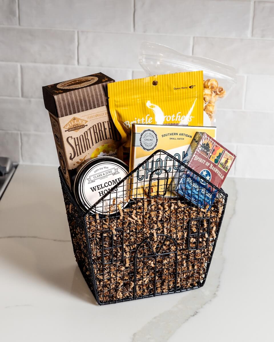 A Taste of the South, Tied with a Bow: High Note Gifts - 7 A gift basket containing shortbread, brittle, crackers, popcorn, a candle, and a postcard sits on a white countertop.
