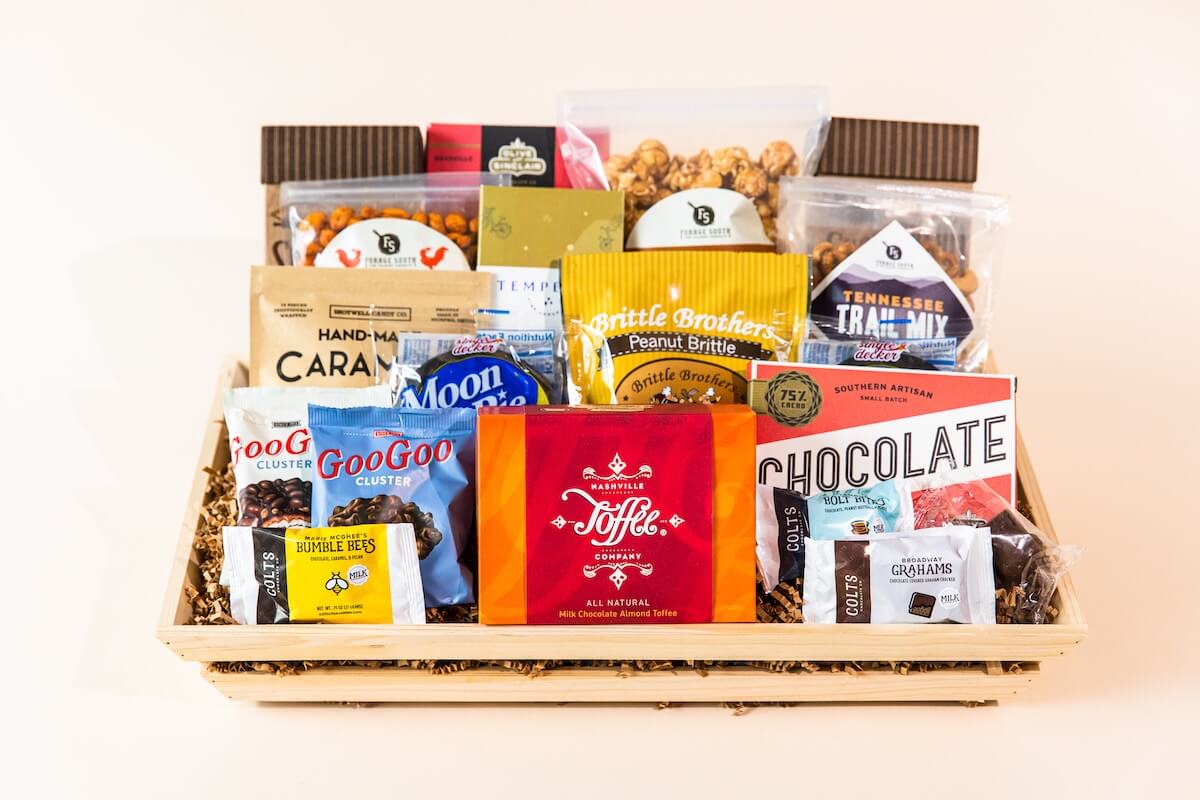 A Taste of the South, Tied with a Bow: High Note Gifts - 2 A wooden tray filled with assorted packaged snacks, including caramel popcorn, chocolate bars, candy, and gourmet treats, arranged neatly against a plain background.