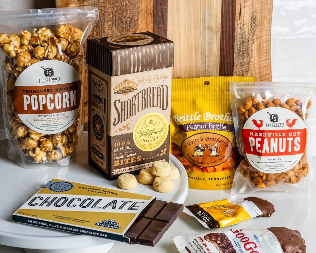 A Taste of the South, Tied with a Bow: High Note Gifts - 3 An assortment of packaged snacks, including caramel popcorn, shortbread bites, peanut brittle, hot peanuts, a chocolate bar, and chocolate-covered candy, arranged on a white surface.