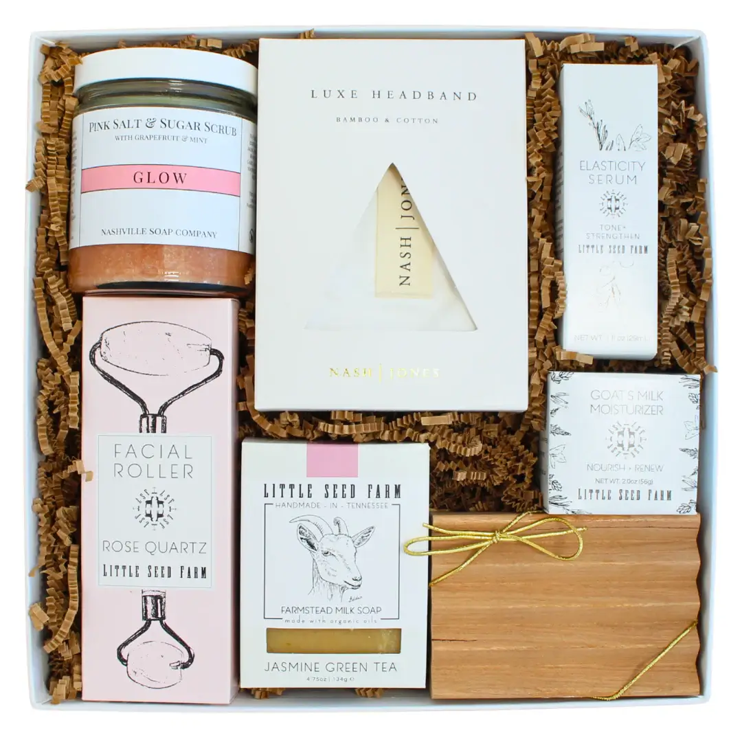 A Taste of the South, Tied with a Bow: High Note Gifts - 6 A gift box from High Note Gifts featuring self-care itemsβpink salt scrub, facial roller, headband, elasticity serum, bar soap, moisturizer, and incense sticksβbeautifully arranged on shredded paper. Perfect for anyone who loves Southern gifts.