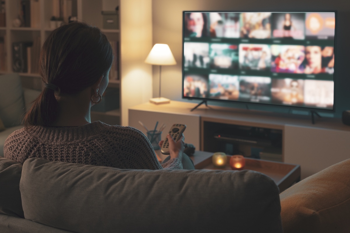 A person sits on a couch in a living room, holding a remote and facing a TV displaying their Southern Gothic-filled Watch List on the streaming service menu.