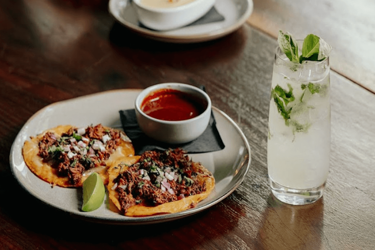Two tacos with shredded meat and diced onions are served with a lime wedge and red sauce on a plate, next to a glass of clear drink with mint leaves.