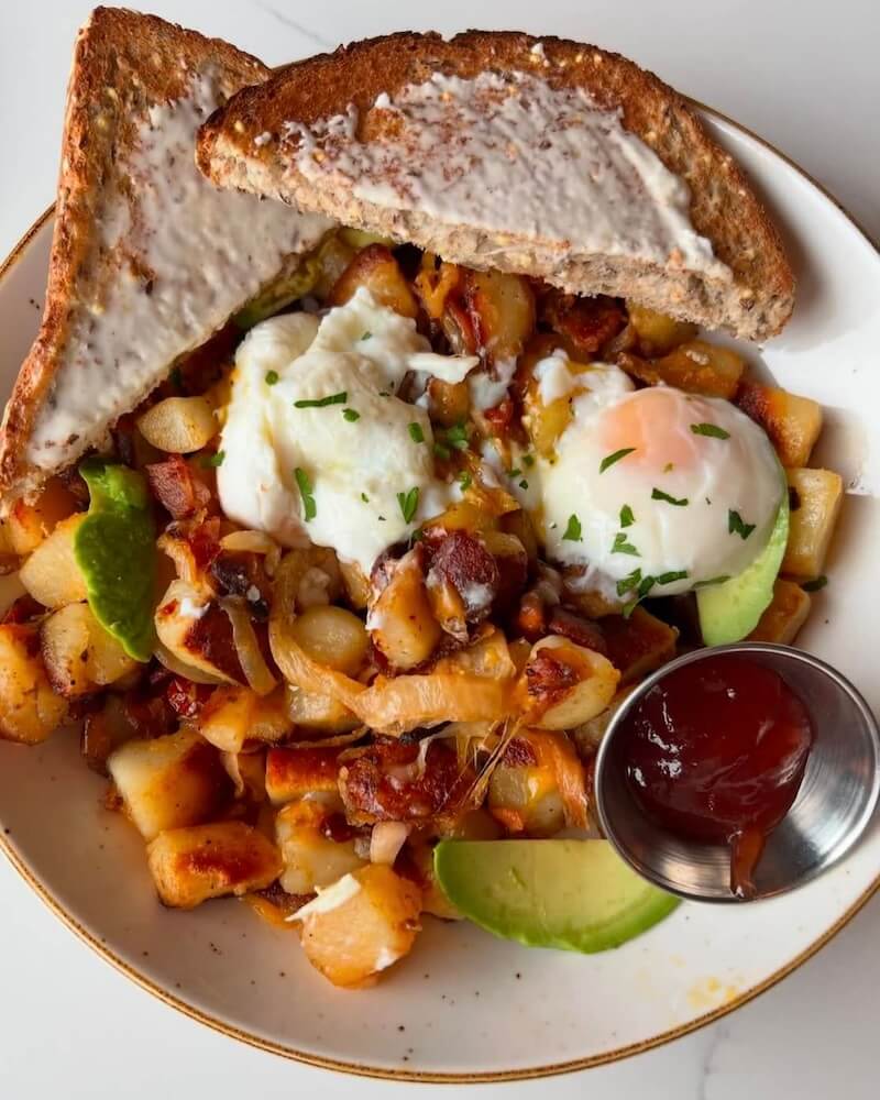 A plate with two slices of toast, two poached eggs, diced potatoes with herbs, avocado slices, and a small cup of ketchup.