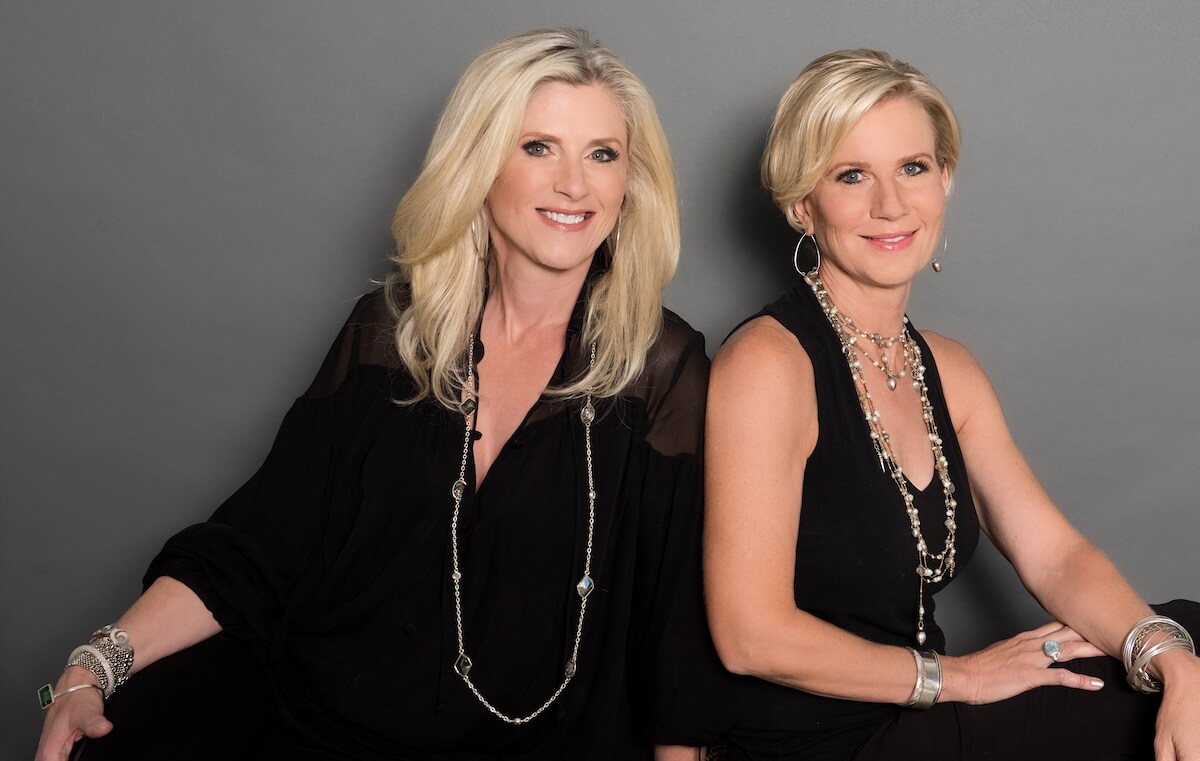 Meet the Duo Behind the Plastic Surgery Center of Nashville - 2 Two women with blonde hair, dressed in black outfits and layered jewelry, sit against a gray background, smiling at the camera.