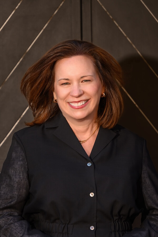 Meet Podcaster Monica Samuels of "Inside the Moms Club" - 2 A woman with shoulder-length brown hair wearing a black button-up top and gold jewelry smiles in front of a dark, geometric-patterned background, radiating confidence as if she's inside the mom's club.