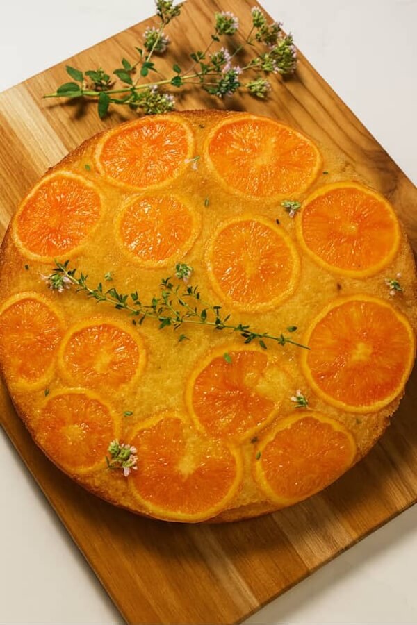 7 New Nashville Restaurants to Try This Month - 2 A round orange upside-down cake topped with orange slices and small sprigs of herbs, placed on a wooden cutting board with extra herb sprigs beside it.