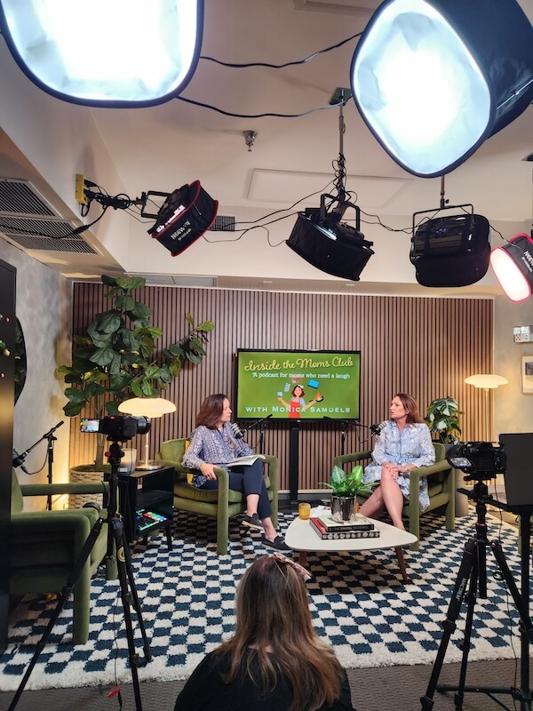 Meet Podcaster Monica Samuels of "Inside the Moms Club" - 3 Two women sit and talk on a set designed for a talk show, with cameras and studio lights visible, and a sign reading “Inside the Mom’s Club” in the background, capturing the lively spirit of Inside the Mom’s Club.
