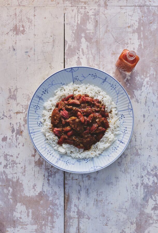 8 Cozy, Cajun-Inspired Recipes for Fat Tuesday - 5 A plate of white rice topped with a stew of red beans and meat, placed on a rustic wooden surface, with a small bottle of hot sauce beside it.
