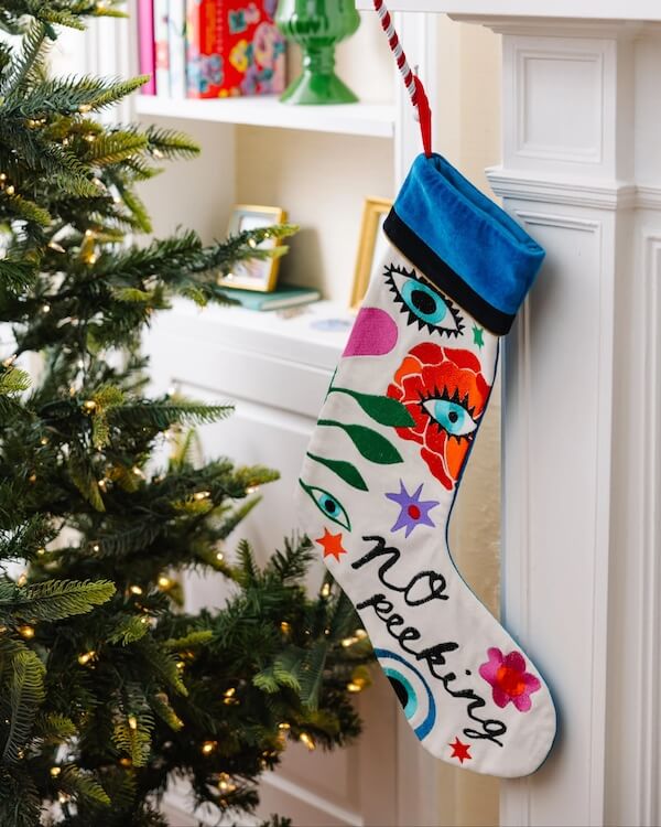 14 Southern Ornament & Stocking Shops We Love - 6 A colorful Christmas stocking with eye and flower designs and the words "NO peeking" hangs on a white mantel near a decorated Christmas tree adorned with southern ornaments.