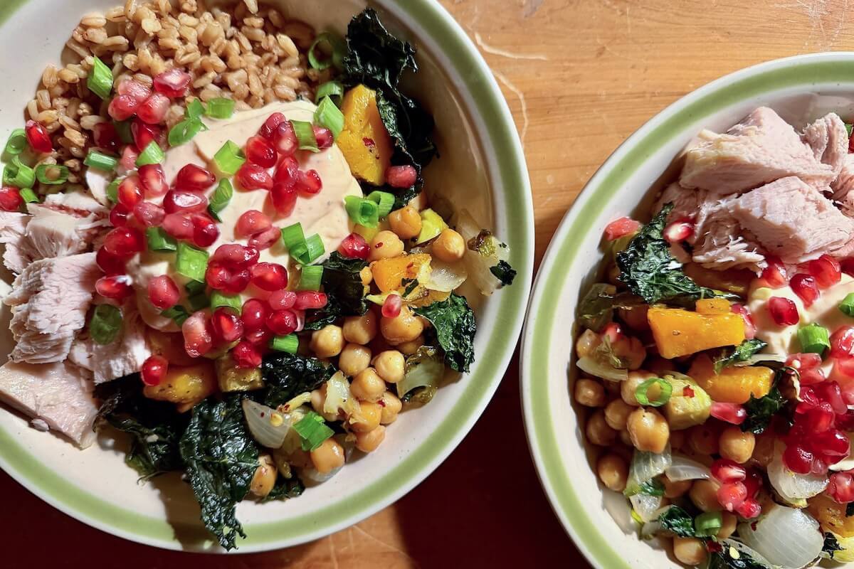 Two bowls of High-Protein Farro Bowl with cooked turkey, chickpeas, roasted vegetables like kale and squash, onions, green onions, pomegranate seeds, and a dollop of sauce, all served on a wooden surface.