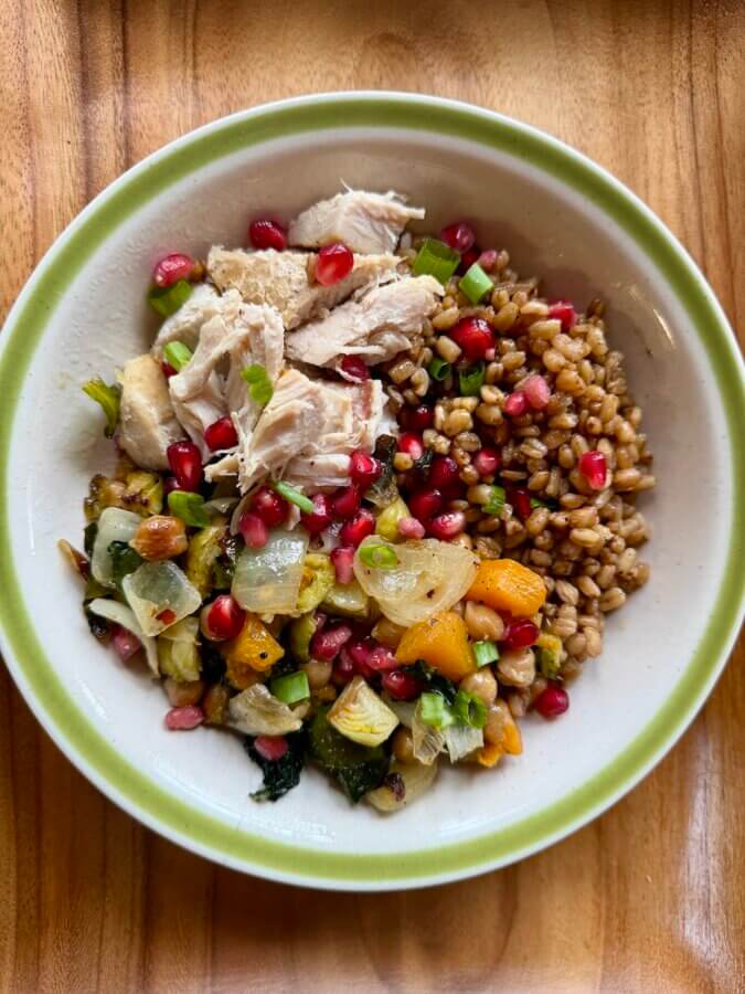 High-Protein Farro Bowl With Roasted Vegetables, Chicken & Yogurt Sauce - 2 A High-Protein Farro Bowl featuring shredded chicken, roasted vegetables, pomegranate seeds, and green onions sits on a wooden surface. Serve with a dollop of yogurt sauce for extra flavor.