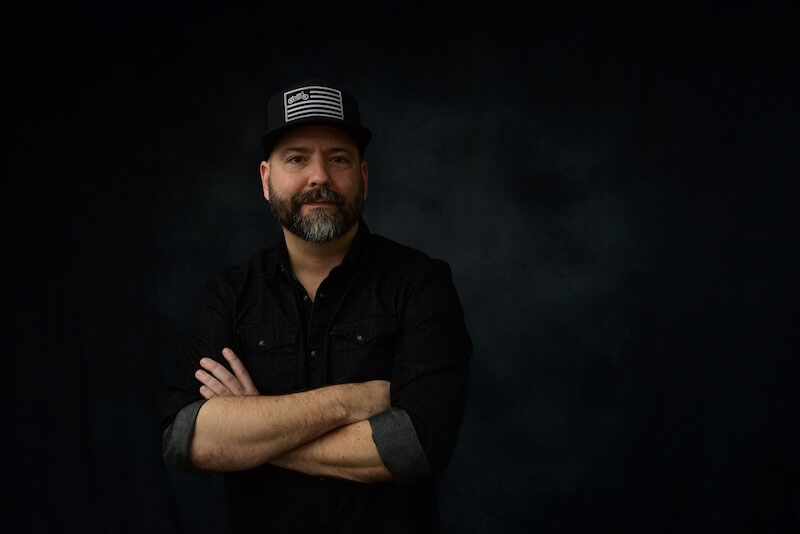 Justin Orton, a man with a beard, wears a black shirt and a hat with a flag design, standing against a dark background with his arms crossed.