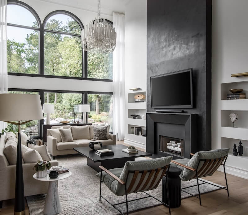 Modern living room with tall windows, neutral-toned furniture, a black fireplace with mounted TV, chandelier, and built-in shelves overlooking a lush green outdoor view.